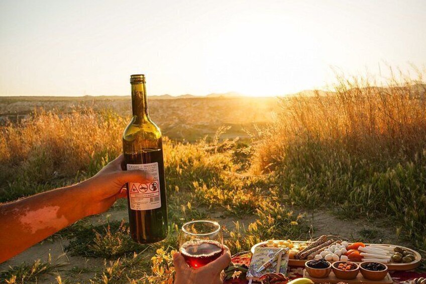 Una botella de vino, una copa y una tabla de picnic con queso, pan y aceitunas en un campo de hierba al atardecer. 2