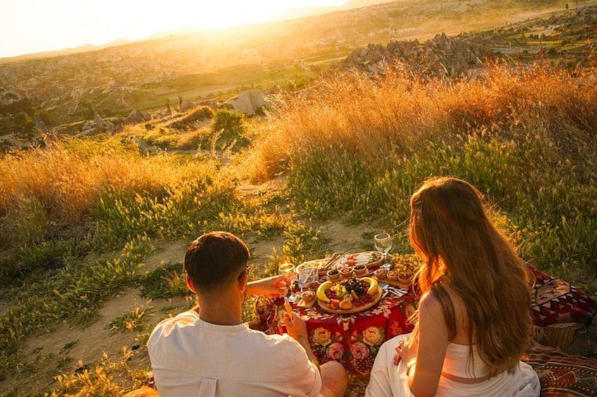 Un couple est assis à une table de pique-nique, devant un repas, et contemple un paysage verdoyant avec un coucher de soleil en arrière-plan. 2