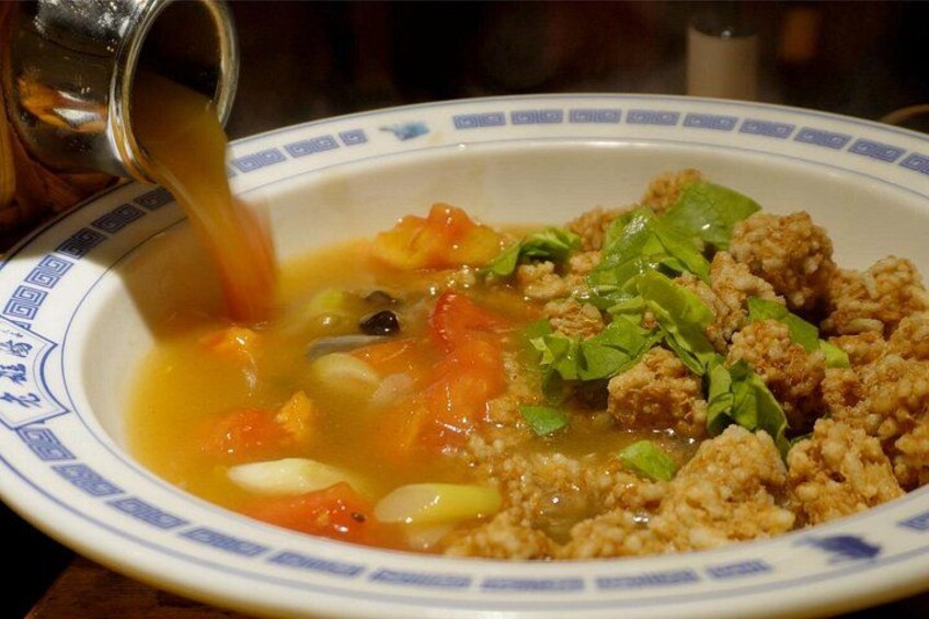A bowl of soup with vegetables and meat, being poured from a pitcher. 3
