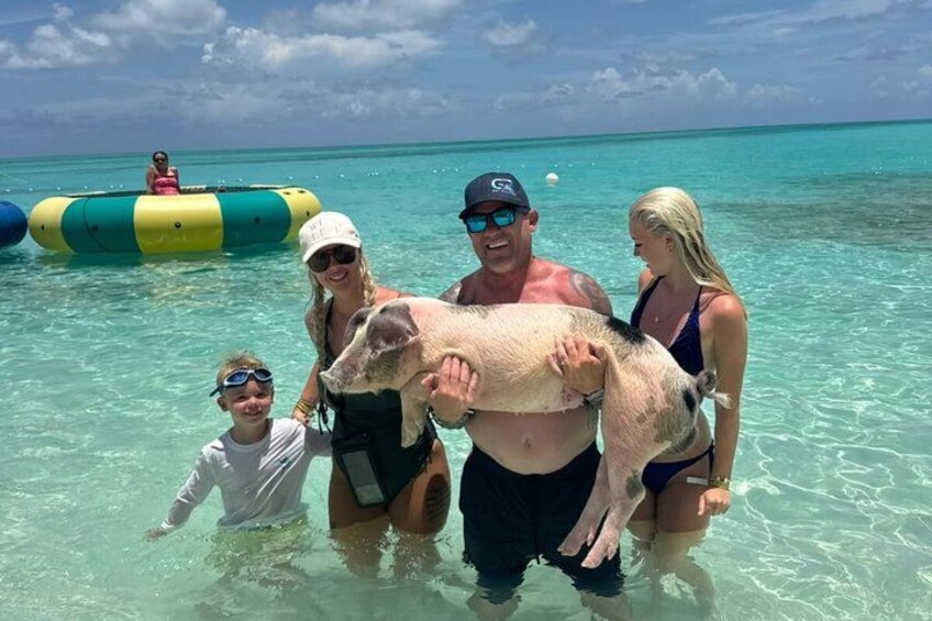 Un groupe de personnes tenant un cochon dans une eau turquoise et limpide, avec un radeau en arrière-plan. 1