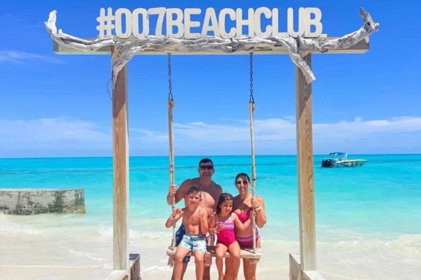 Un columpio de madera con el hashtag #007BEACHCLUB, un océano azul cristalino y un barco a lo lejos. 4