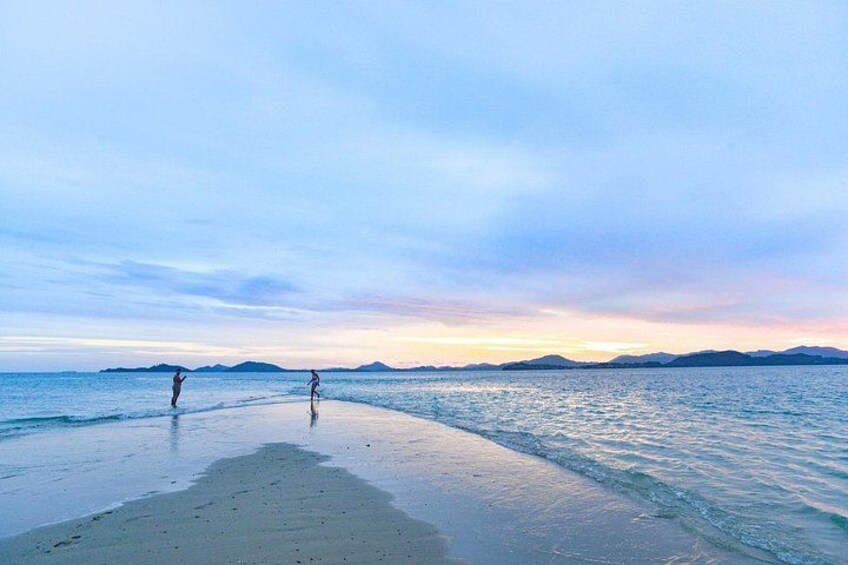 A beach with two people walking along the shore, a calm sea, and a sunset in the background. 3