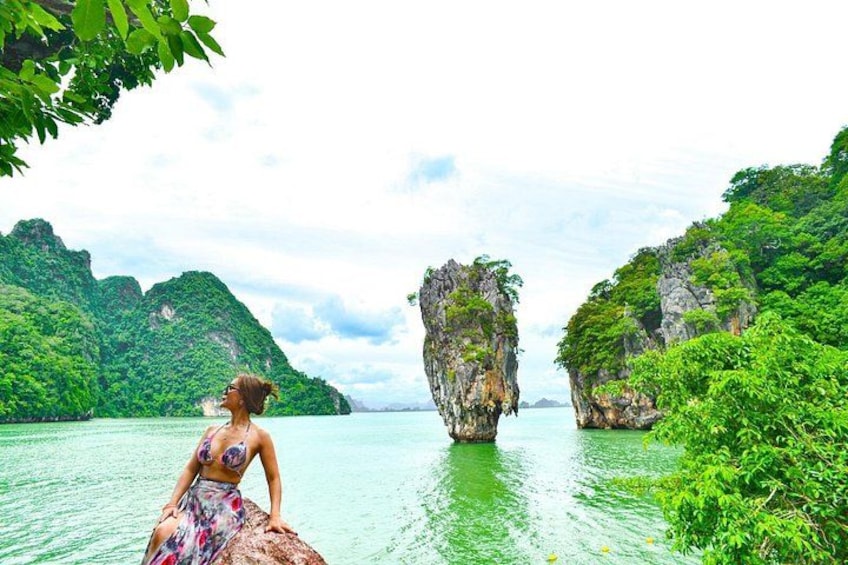 A woman sitting on a rock by a body of water with green foliage and rock formations in the background. 1