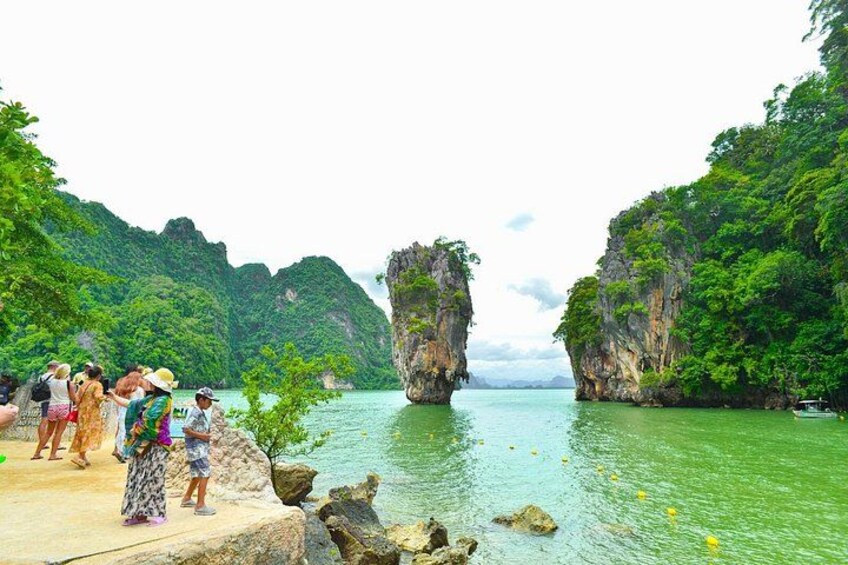 A group of people on a rocky shore, a large rock formation in the water, and lush green hills in the background. 2