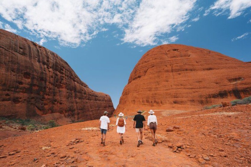 A group of people walking on a red dirt path between two large red rock formations under a clear sky. 5