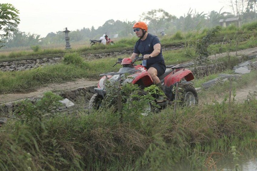 A person riding a red ATV through a rural landscape with water channels and vegetation. 5