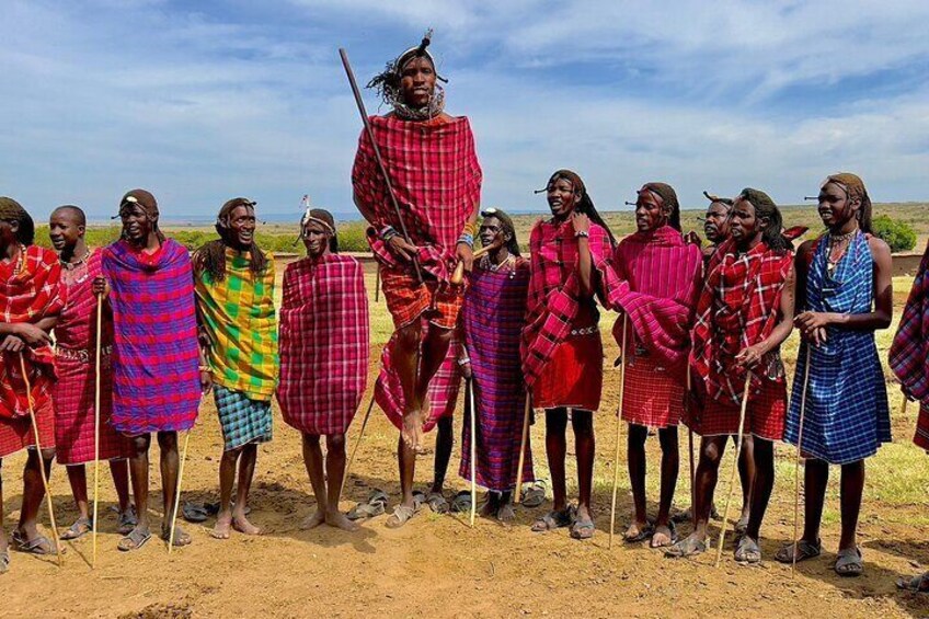 A group of people in traditional attire standing on a dirt ground with a clear sky in the background. 2