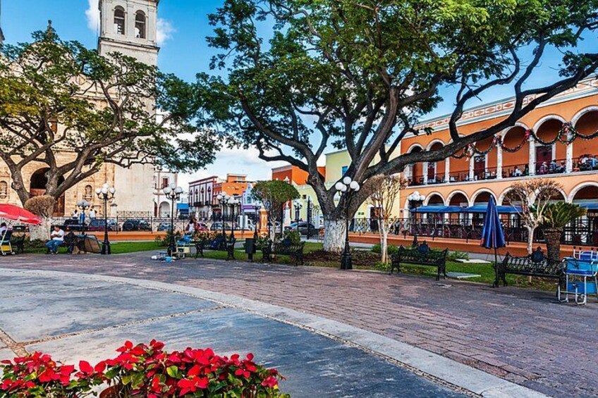 Una plaza con un árbol grande, un edificio con arcos y una torre del reloj. 1