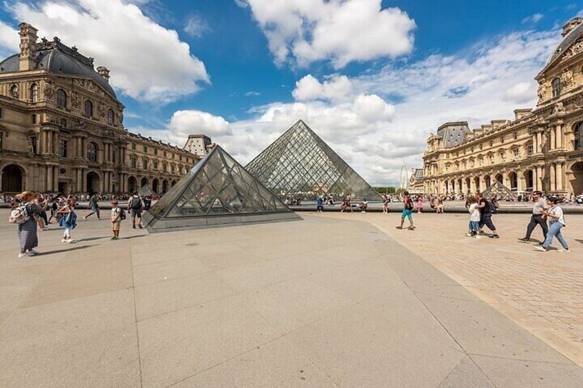 The Louvre Pyramid, a large glass pyramid, surrounded by historic buildings and a paved plaza with people walking around. 2
