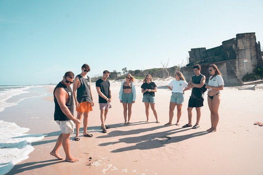A group of people standing on a sandy beach with ocean waves in the background and a large concrete structure to the right. 1