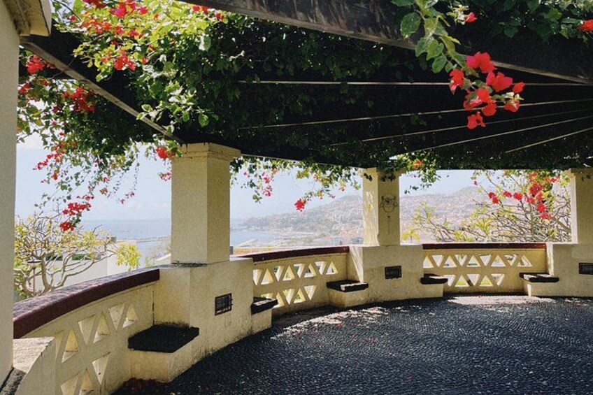 A covered outdoor area with a view of the sea, featuring a pergola with red flowers, a tiled floor, and a decorative wall with geometric patterns. 5