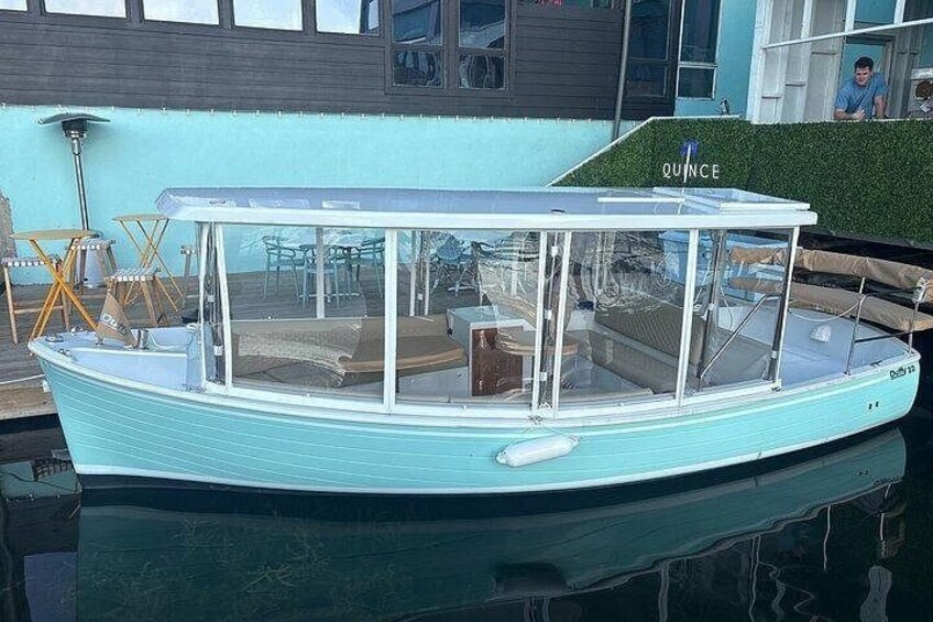 A light blue boat named 'QUINCE' docked at a marina with a grassy area and a building in the background. 2