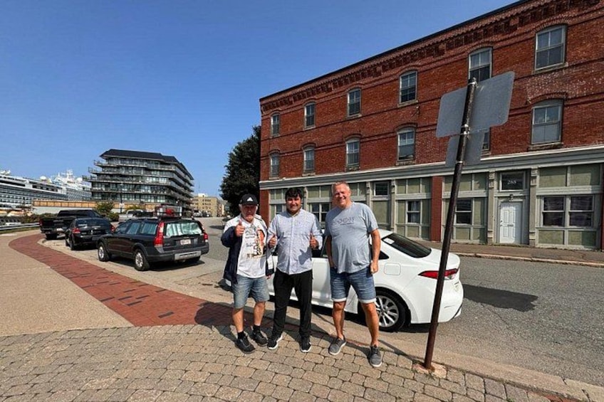 Three people standing on a sidewalk next to a white car, with a brick building and a modern structure in the background. 3