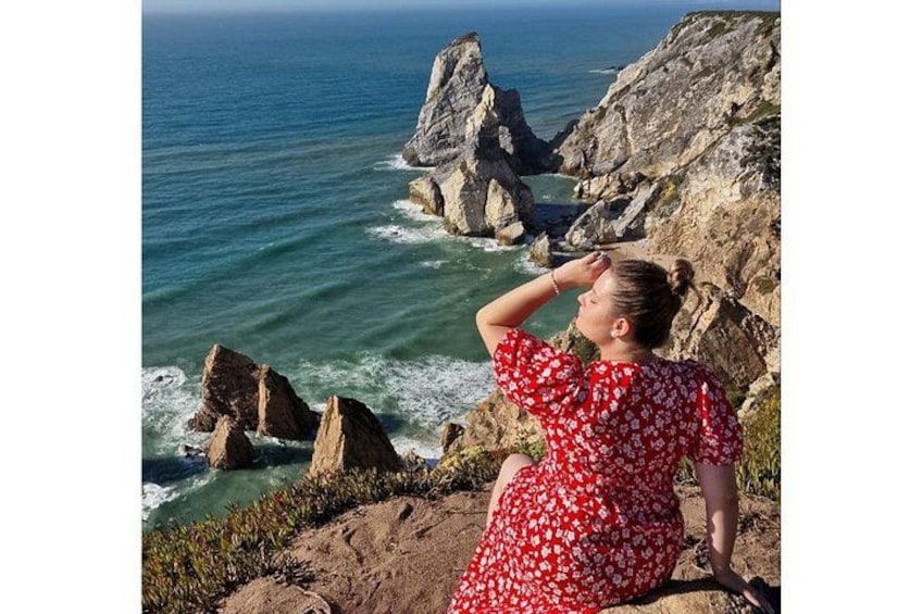 A woman in a red dress with white patterns, standing on a rocky cliff overlooking the ocean. 2