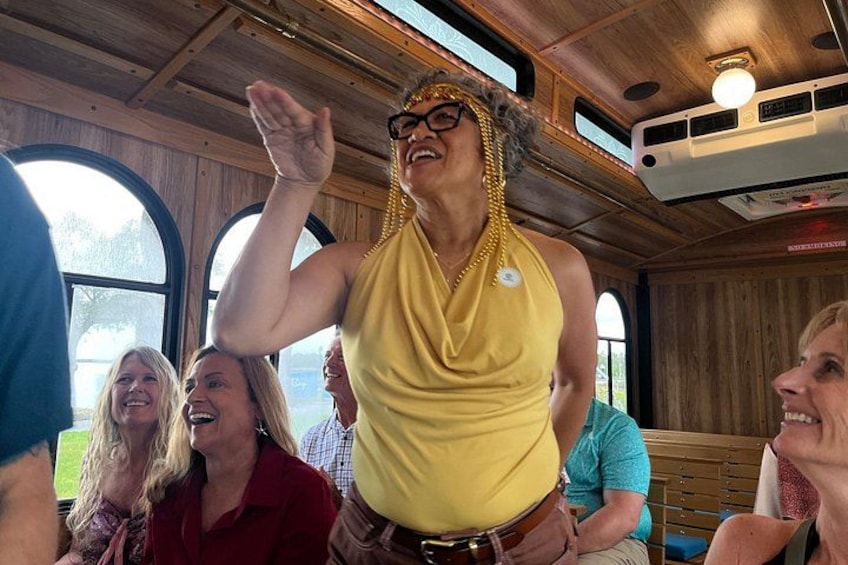 A person in a yellow sleeveless top and gold jewelry, seated in a vehicle with wooden interior and windows. 5