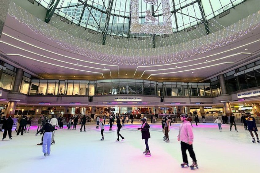An indoor ice skating rink with people skating, a glass ceiling, and shops in the background. 3