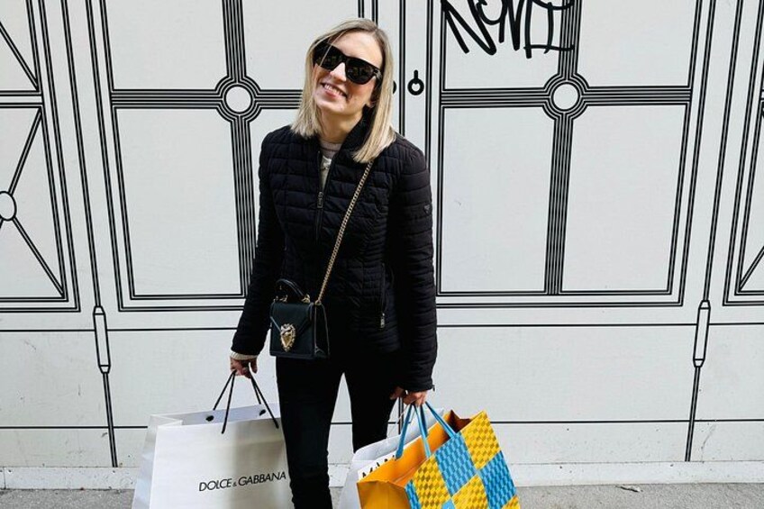 Una persona frente a una pared blanca con patrones geométricos negros, sosteniendo una chaqueta negra y un bolso estampado en amarillo y azul. 1