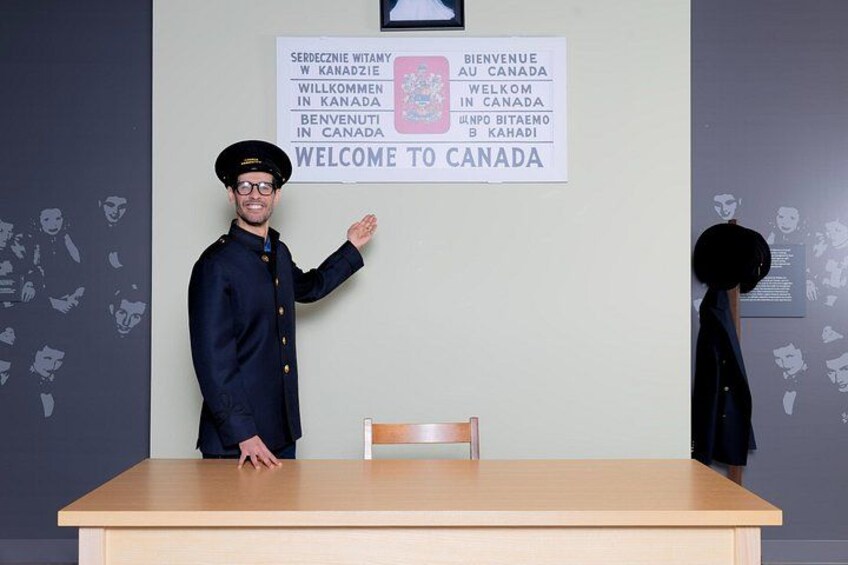 Una persona con uniforme de pie en un mostrador con un cartel que dice 'WELCOME TO CANADA' en varios idiomas. 4