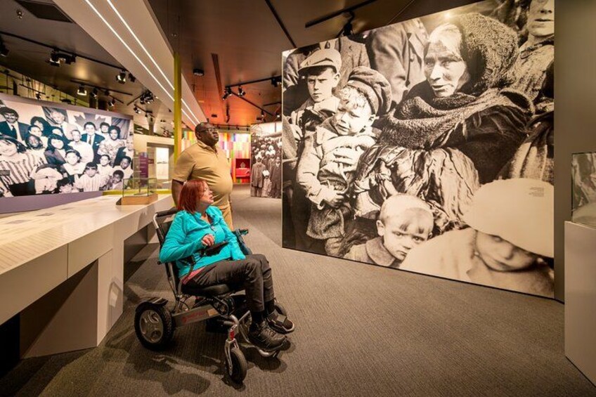 Et museumsinteriør med en køre­stol, et stort sort-hvidt fotografi og en gruppe mennesker. 2