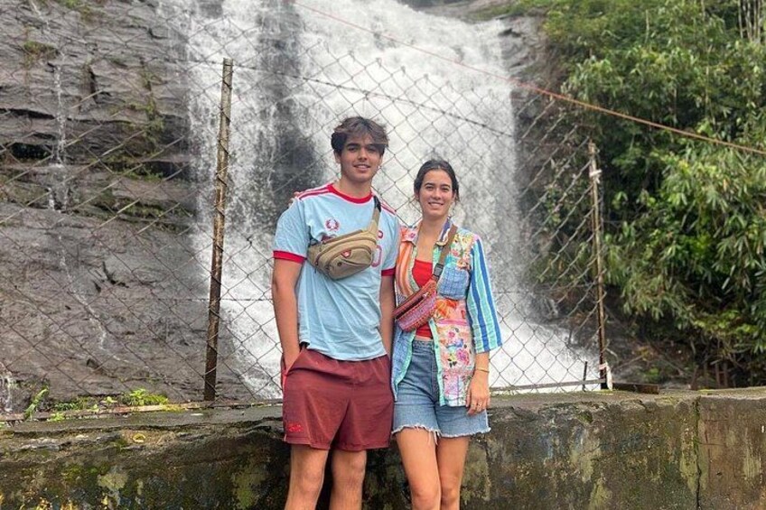 Two people standing in front of a waterfall with a rocky background. 1