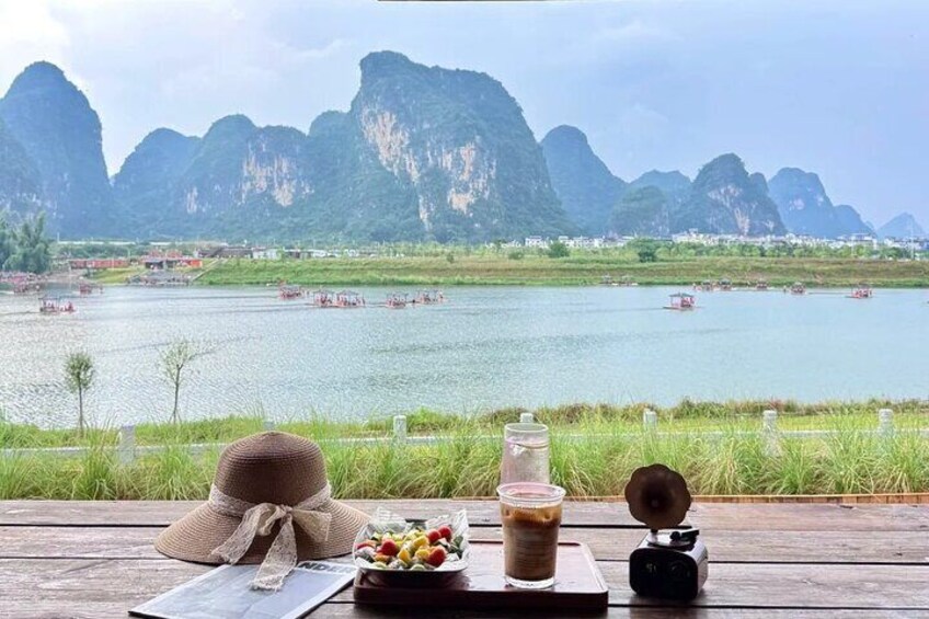 A wooden table with a hat, a glass of iced coffee, and a camera. In the background, there is a river with boats and a mountainous landscape. 2