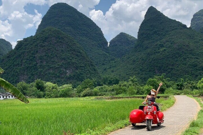 A person on a red motorcycle with large sidecars, riding on a paved path through a green field with mountains in the background. 3