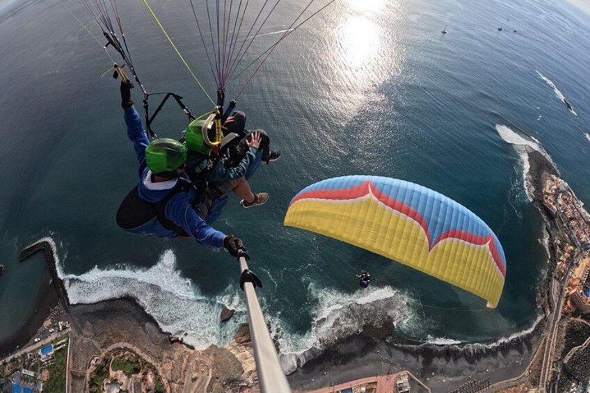 Een persoon paraglidet boven een kustgebied met een kleurrijke parachute, golven die op het strand breken, en een verre kustlijn. 2