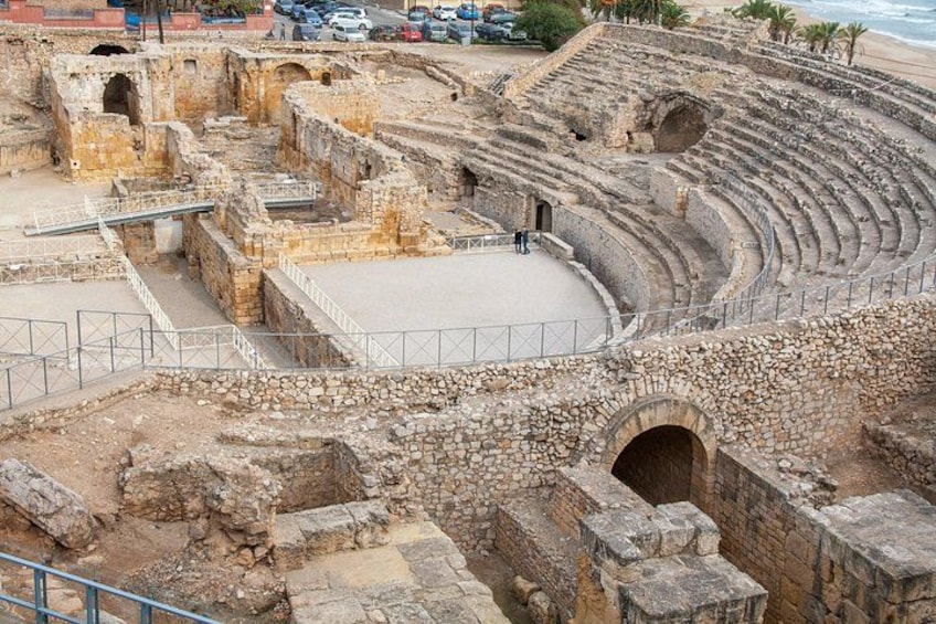 Ein antikes Amphitheater mit Steinsitzen und einer zentralen Bühne, umgeben von Ruinen und modernen Gebäuden. 4