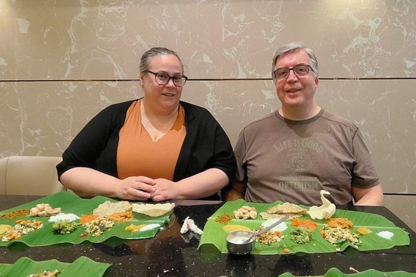 Two people seated at a table with food arranged on banana leaves. 1
