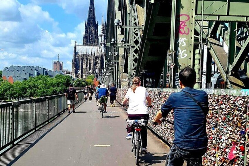 A bridge with cyclists and pedestrians, a gothic cathedral in the background, and a wall covered in small objects. 1
