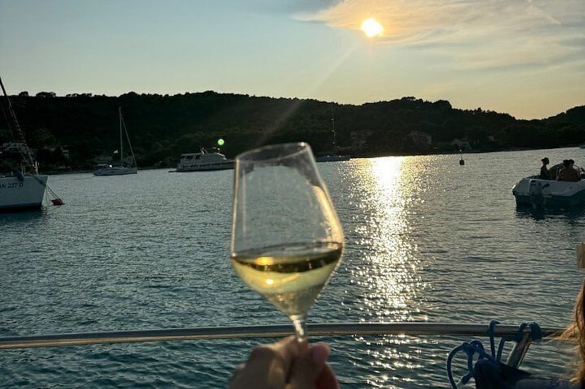 A glass of white wine held by a person, a boat with people on it, and a sunset over a body of water with boats and a forested hill in the background. 2