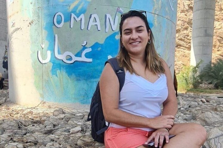 Una persona seduta di fronte a un cartello con la scritta 'OMAN' e caratteri arabi. 1