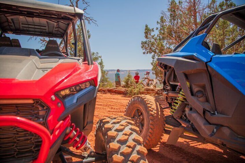 A red and a blue off-road vehicle parked on a dirt road with a person in red standing in the background. 2