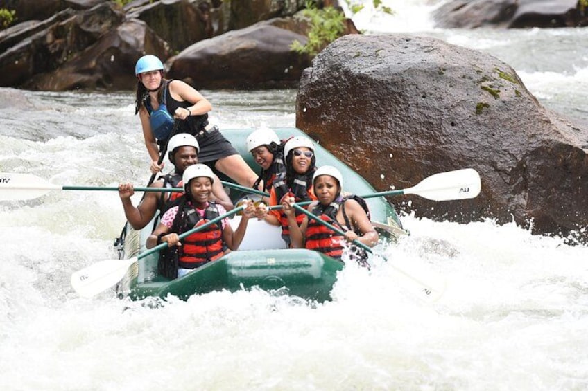 A group of people in a green raft navigating a river with rapids, surrounded by large rocks and trees. 1