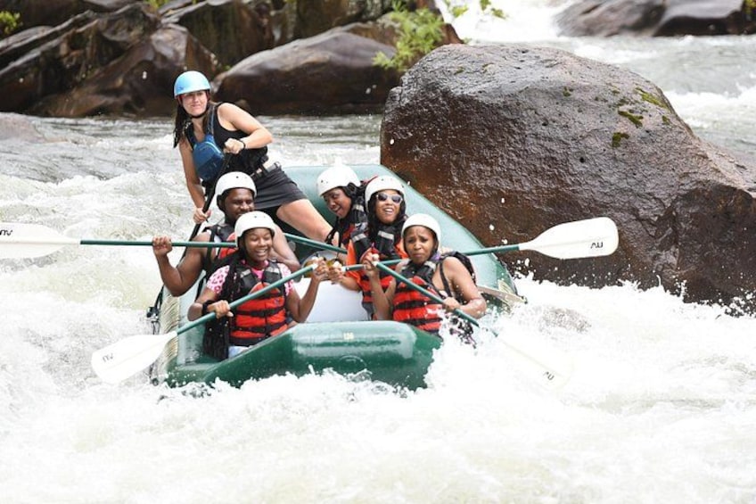 A group of people in a green raft navigating a river with rapids, surrounded by large rocks and trees. 1