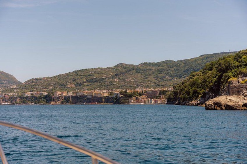 A coastal town with buildings along the shore, a rocky outcrop, and a hilly landscape in the background. 4