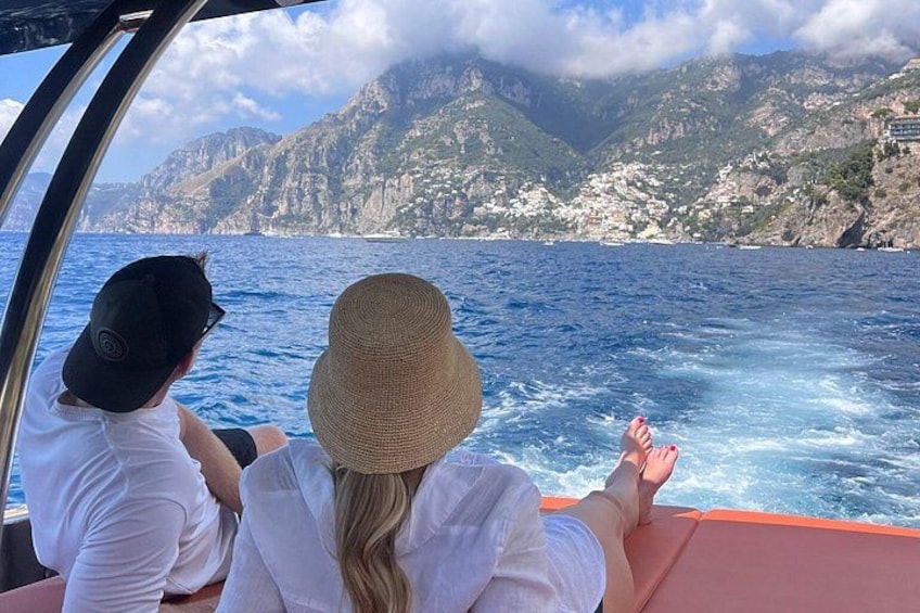 Two people on a boat, one wearing a hat, with a mountainous coastline in the background. 1