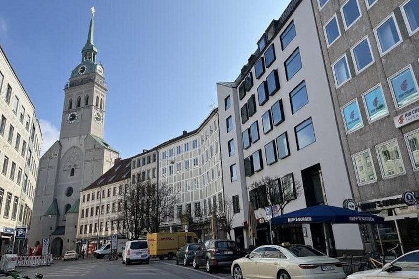 En gade i byen med et markant klokketårn, moderne bygninger og parkerede biler. 3