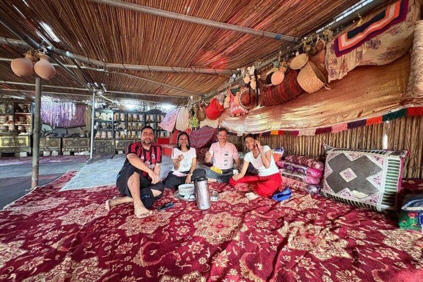 A group of people sitting on a patterned carpet under a thatched roof, surrounded by textiles and pottery. 2