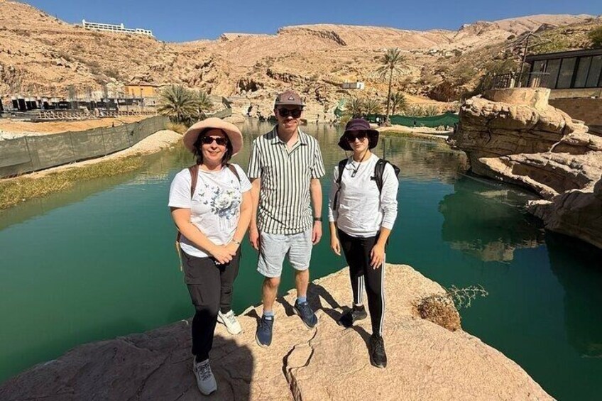 Three people standing by a body of water with rocky terrain and palm trees in the background. 1