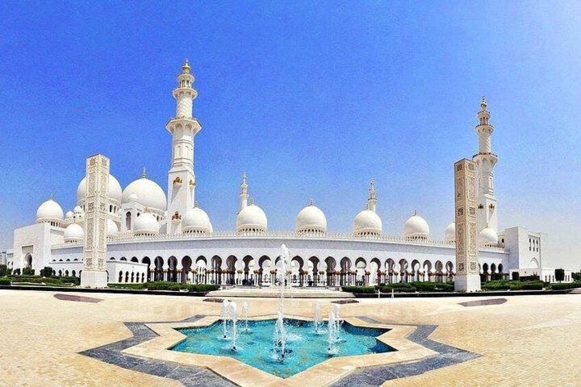 A large white mosque with multiple domes and minarets, a central fountain, and a patterned courtyard. 2