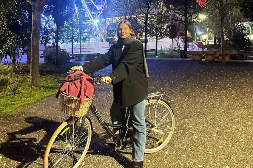 A person standing next to a bicycle with a basket containing a red jacket, in a park with trees and a Ferris wheel in the background. 4
