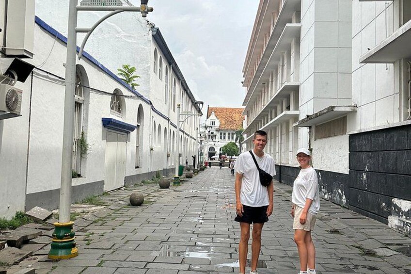 A narrow street with white buildings, a man and a woman standing, and a distant church. 2