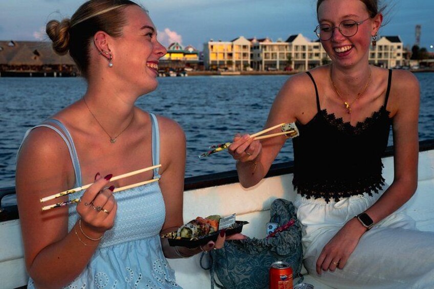 Two people on a boat, holding chopsticks, with a body of water and buildings in the background. 2