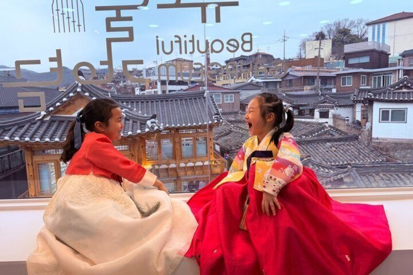 Two individuals in traditional Korean hanbok sitting on a ledge with a backdrop of Korean rooftops. 1