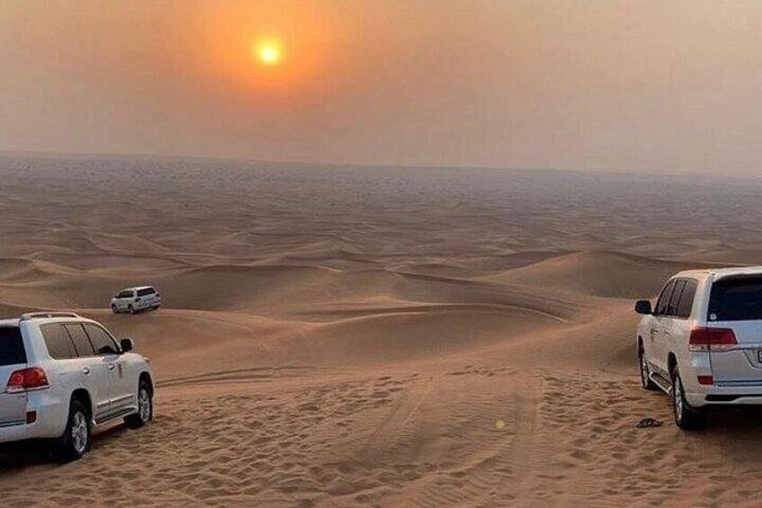 A desert landscape with two cars parked on sand dunes, a clear sky, and the sun setting in the background. 3
