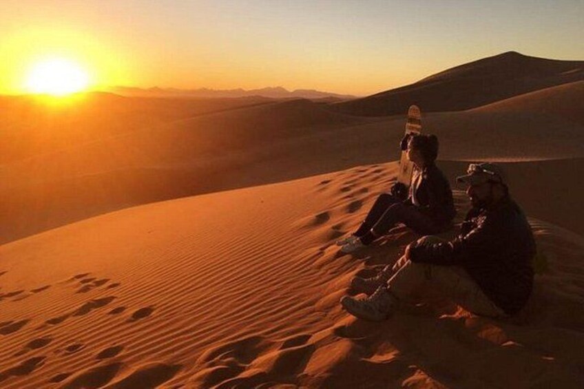Two people sitting on a sand dune with a sunset in the background. 2