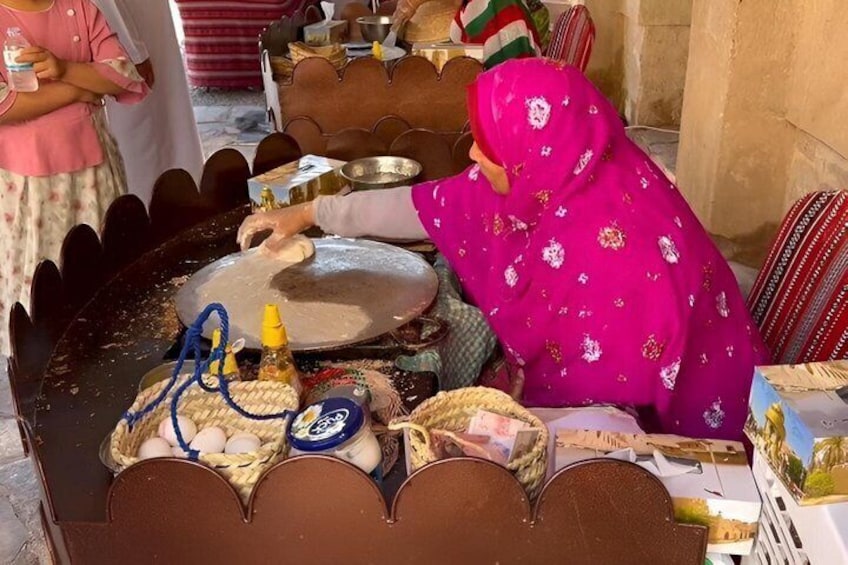 Una persona con un chal rosa estampado preparando comida en una plancha redonda grande, con una cesta de huevos y una botella de aceite cerca. 5