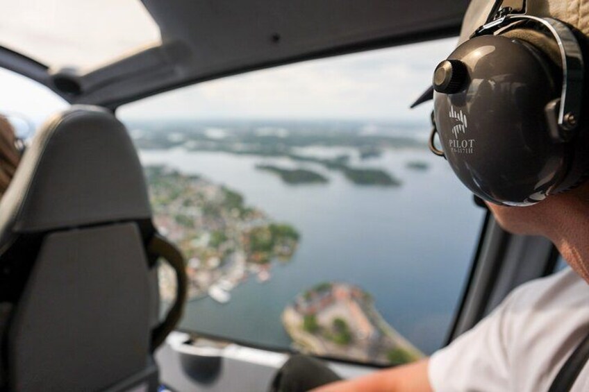 A pilot wearing a headset with 'PILOT' visible, inside a helicopter cockpit with a view of a coastal landscape. 2