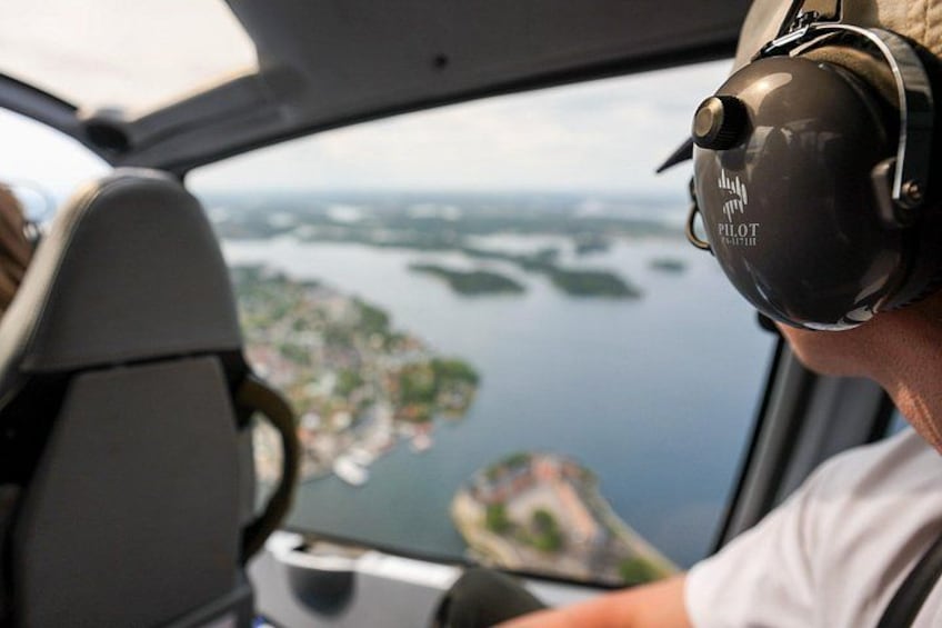 A pilot wearing a headset with the word 'PILOT' visible, seated in a cockpit with a view of a coastal landscape through the windshield. 2